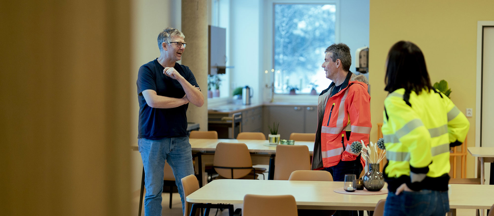 Tre personer som står inne i et rom og prater sammen. Foto.