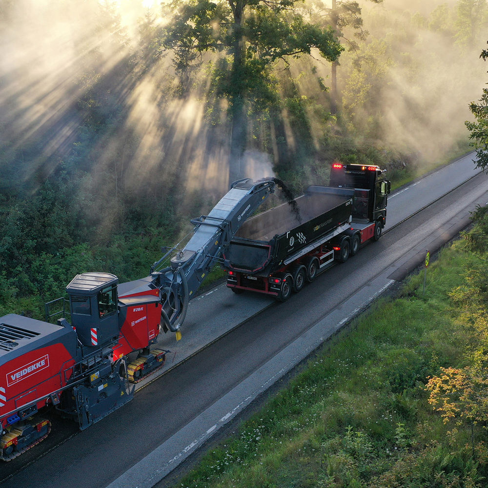 Asfaltfres og lastebil på vei med solstråler gjennom skog. Foto.