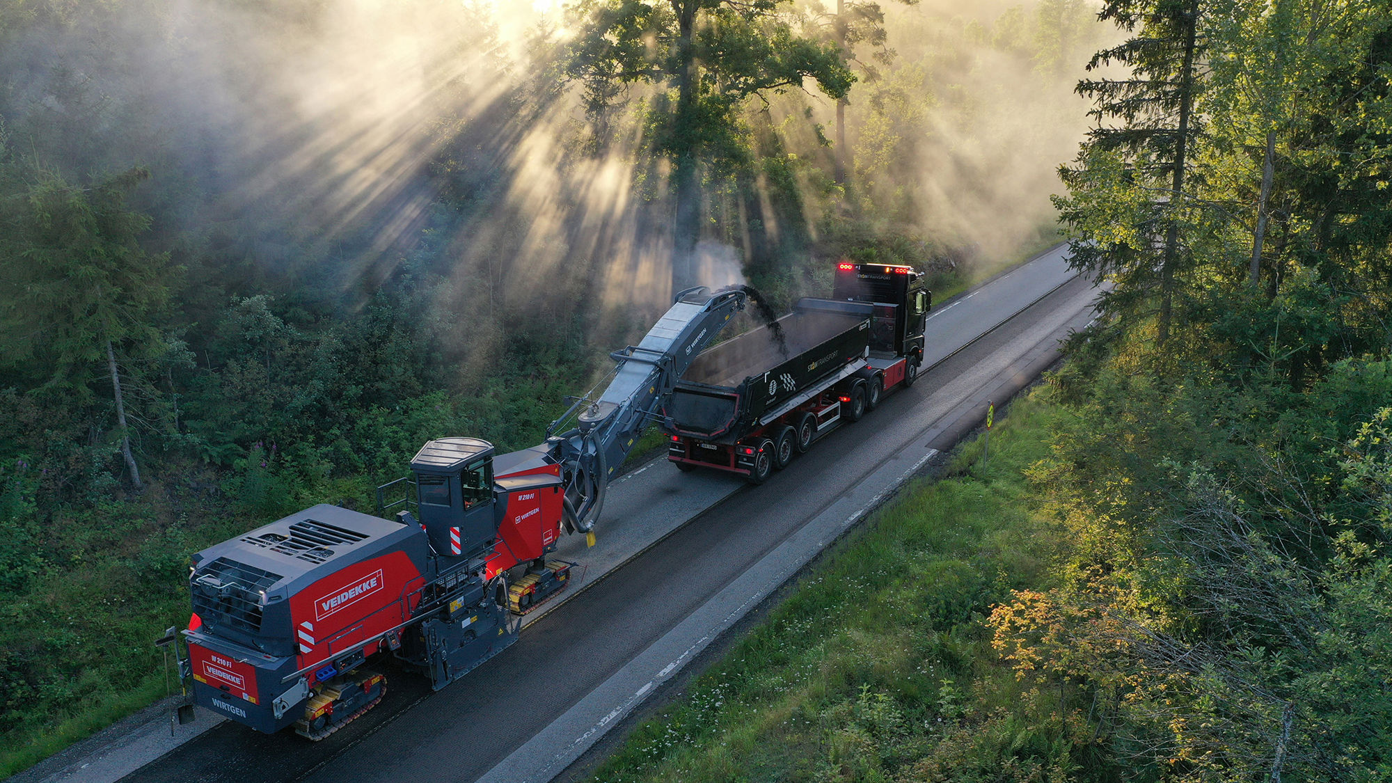 Asfaltfres og lastebil på vei med solstråler gjennom skog. Foto.